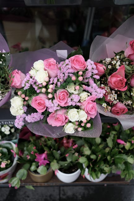 A floral arrangement featuring a bouquet of fresh pink roses, white and pink spray carnations, and clusters of delicate pink astilbe, all arranged with lush green foliage. The bouquet is wrapped in tr