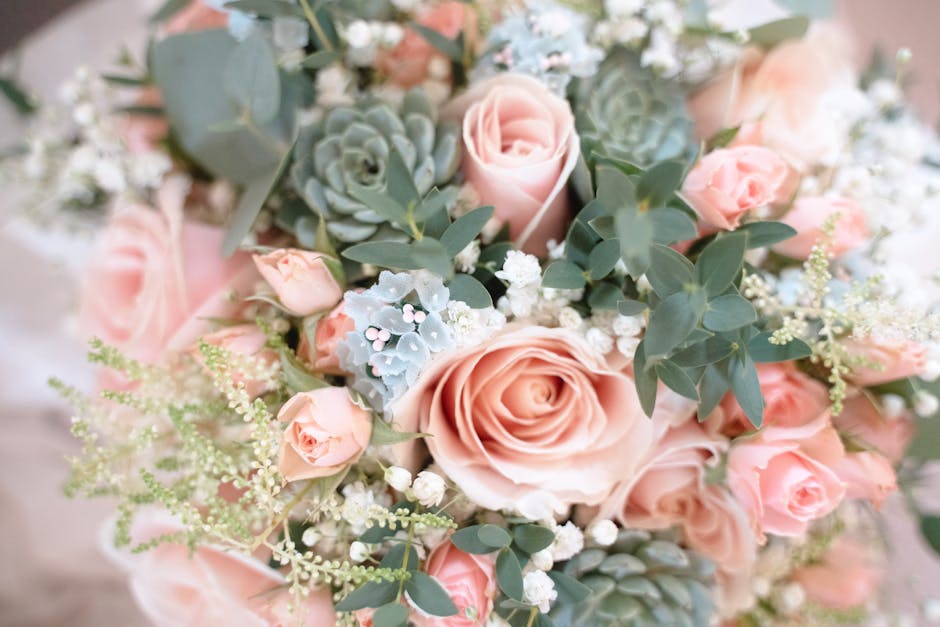 A close-up view of a floral bouquet featuring large, soft pink roses with delicate, layered petals, surrounded by small pink spray roses, silvery-green eucalyptus leaves, and clusters of tiny white ba