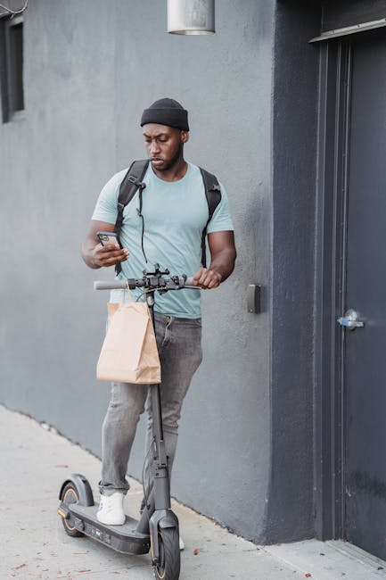 A young man with dark skin, wearing a black beanie, light gray T-shirt, and dark jeans, stands on a sidewalk next to a gray building, looking at his smartphone. He holds a brown paper bag in his left