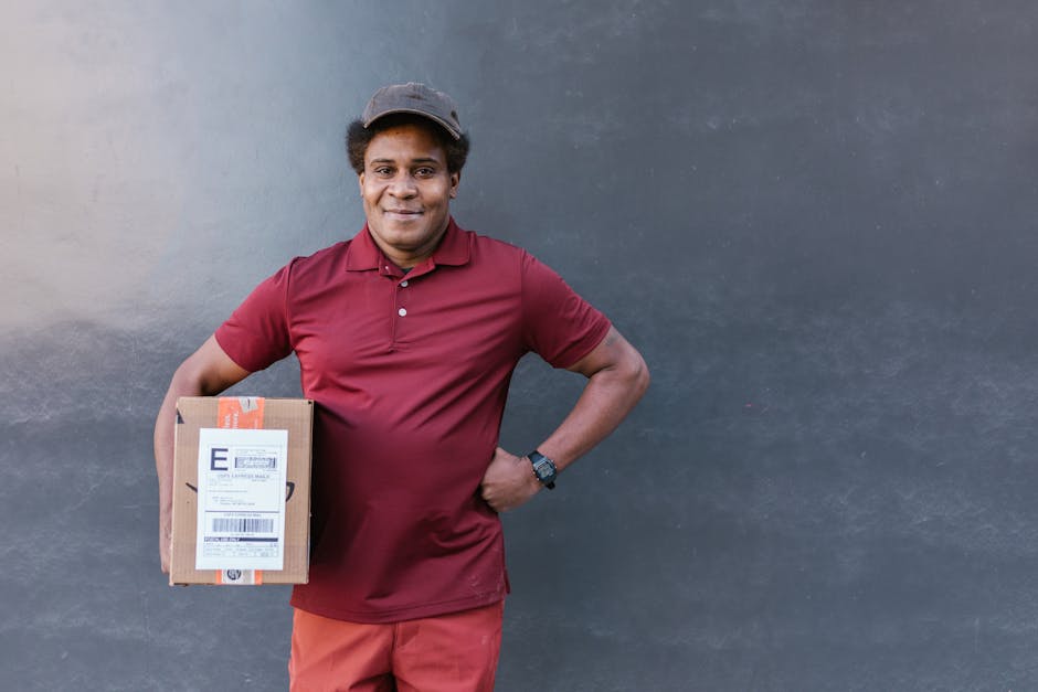 A delivery person with dark skin and curly hair, wearing a maroon polo shirt, maroon trousers, and a dark cap, stands against a plain, dark-colored wall. He is smiling and holding a small cardboard bo
