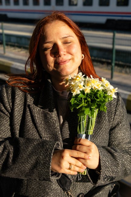 A woman with red hair, smiling gently with her eyes closed, holds a fresh bouquet of yellow and white daffodils with green foliage, wrapped in clear glass, standing outdoors near a railway station pla